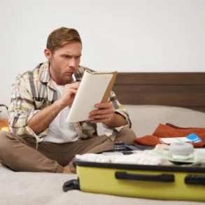 Man sitting cross-legged on bed, checking a list while packing a yellow suitcase with clothes and toiletries.