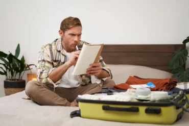 Man sitting cross-legged on bed, checking a list while packing a yellow suitcase with clothes and toiletries.