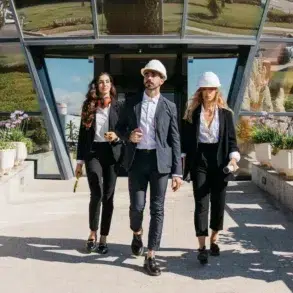 Three business professionals in suits and hard hats confidently walk out of a modern building, surrounded by potted plants on a sunny day.