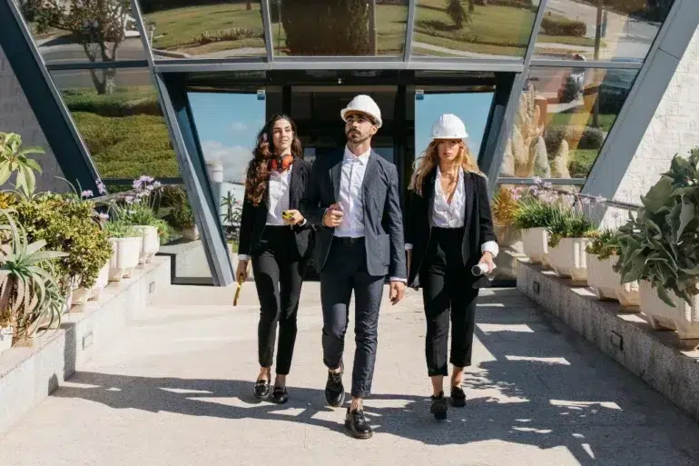 Three business professionals in suits and hard hats confidently walk out of a modern building, surrounded by potted plants on a sunny day.