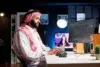 A man in traditional Middle Eastern attire sits at a desk, focused on a computer screen displaying code. The room is dimly lit with a modern, tech-savvy ambiance.