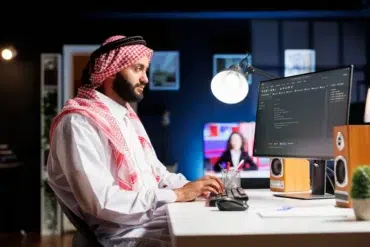 A man in traditional Middle Eastern attire sits at a desk, focused on a computer screen displaying code. The room is dimly lit with a modern, tech-savvy ambiance.