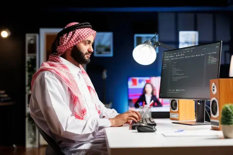 A man in traditional Middle Eastern attire sits at a desk, focused on a computer screen displaying code. The room is dimly lit with a modern, tech-savvy ambiance.