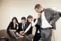 Four people in business attire are gathered around a laptop at a conference table, looking intently at the screen.