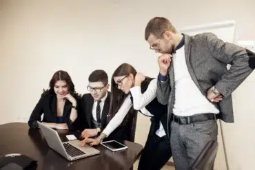 Four people in business attire are gathered around a laptop at a conference table, looking intently at the screen.