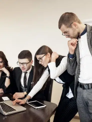 Four people in business attire are gathered around a laptop at a conference table, looking intently at the screen.