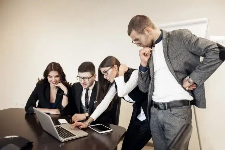 Four people in business attire are gathered around a laptop at a conference table, looking intently at the screen.