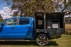 Blue pickup truck with black utility bed featuring open storage compartments under a large tree in a grassy area.