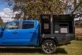Blue pickup truck with black utility bed featuring open storage compartments under a large tree in a grassy area.