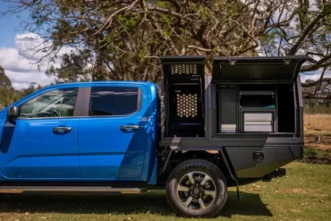 Blue pickup truck with black utility bed featuring open storage compartments under a large tree in a grassy area.
