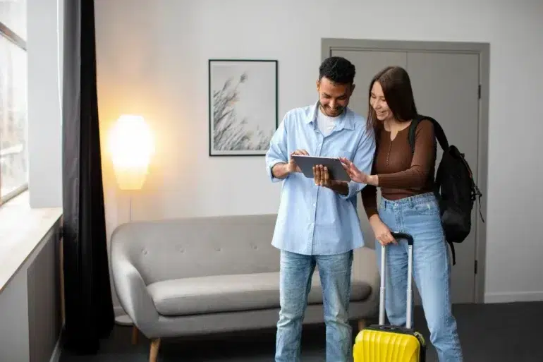 A man and woman stand in a room looking at a tablet, with a suitcase and backpack ready for travel.