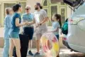 A multi-generational family stands near a car with its trunk open, preparing for a trip with luggage and a colorful inflatable ring.