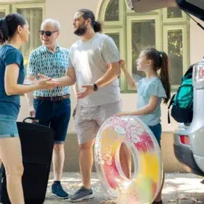 A multi-generational family stands near a car with its trunk open, preparing for a trip with luggage and a colorful inflatable ring.