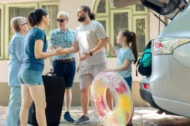 A multi-generational family stands near a car with its trunk open, preparing for a trip with luggage and a colorful inflatable ring.