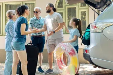 A multi-generational family stands near a car with its trunk open, preparing for a trip with luggage and a colorful inflatable ring.