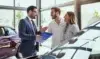 A car salesperson in a suit talks to a smiling couple in a bright dealership. They stand near cars, holding a clipboard, creating a welcoming atmosphere.