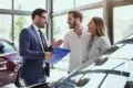 A car salesperson in a suit talks to a smiling couple in a bright dealership. They stand near cars, holding a clipboard, creating a welcoming atmosphere.