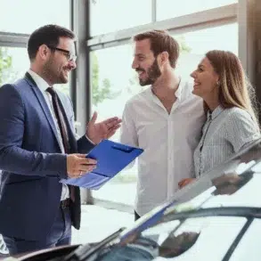 A car salesperson in a suit talks to a smiling couple in a bright dealership. They stand near cars, holding a clipboard, creating a welcoming atmosphere.