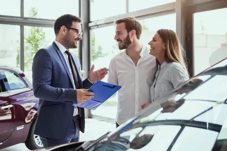 A car salesperson in a suit talks to a smiling couple in a bright dealership. They stand near cars, holding a clipboard, creating a welcoming atmosphere.