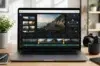 A laptop displaying a video editing software interface. The screen shows a coastal road video clip. Surrounding items include a camera, notebook, and plant, conveying a creative workspace vibe.