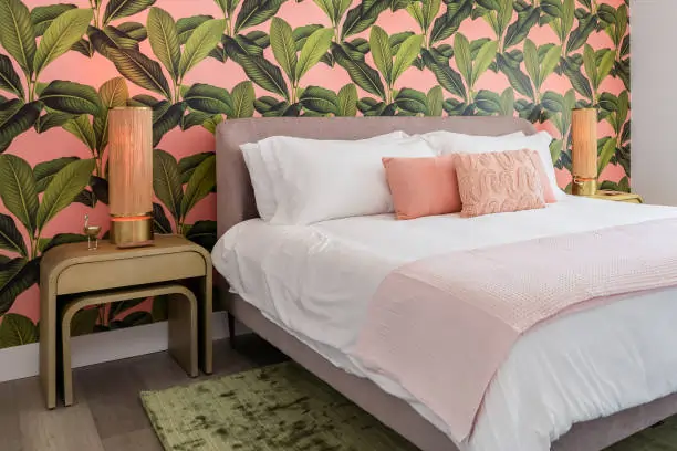 Bedroom with a tropical theme, featuring a bold pink and green leaf wallpaper. Bed with white linens, pink pillows, and elegant side lamps create a cozy ambiance.