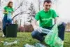 Two volunteers in green shirts collect plastic bottles from a grassy park area.
