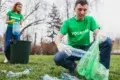 Two volunteers in green shirts collect plastic bottles from a grassy park area.