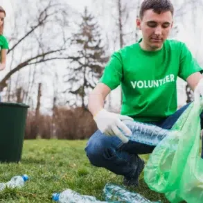 Two volunteers in green shirts collect plastic bottles from a grassy park area.