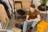 Man using a smartphone sits on the floor surrounded by shopping bags, boxes, and clothes on a rack.