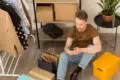 Man using a smartphone sits on the floor surrounded by shopping bags, boxes, and clothes on a rack.