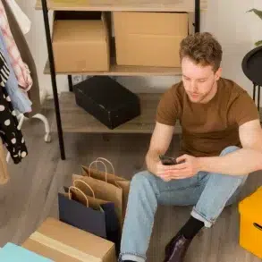 Man using a smartphone sits on the floor surrounded by shopping bags, boxes, and clothes on a rack.
