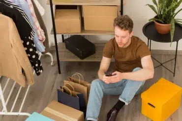 Man using a smartphone sits on the floor surrounded by shopping bags, boxes, and clothes on a rack.