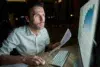 Person in white shirt working late at night on a computer, holding a printed document with charts visible on the monitor.
