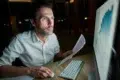 Person in white shirt working late at night on a computer, holding a printed document with charts visible on the monitor.