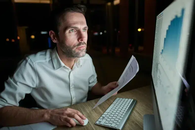 Person in white shirt working late at night on a computer, holding a printed document with charts visible on the monitor.