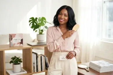 A Black woman in a pink silk shirt and beige pants brushes her dark, wavy hair with a wooden brush while holding a Wigfever business card.