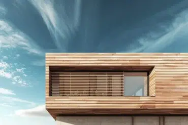 Modern wooden house facade with horizontal slats over a large window under a partly cloudy sky.
