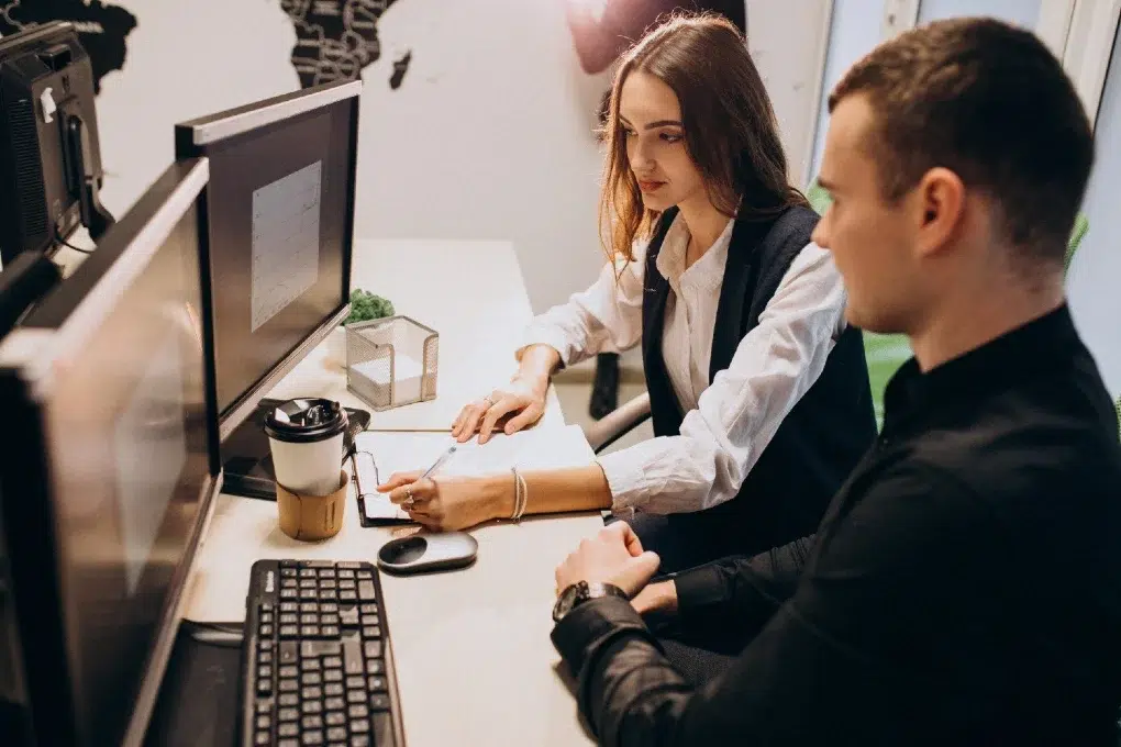 A man and a woman collaborate at a desk with dual monitors, a coffee cup, and documents. The setting is professional and focused.
