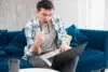 Man in a casual plaid shirt sits on a blue couch, looking at a laptop with a focused expression, holding glasses in one hand. A coffee cup is on a table nearby.