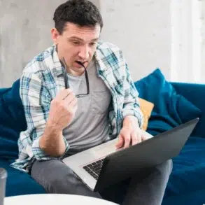 Man in a casual plaid shirt sits on a blue couch, looking at a laptop with a focused expression, holding glasses in one hand. A coffee cup is on a table nearby.