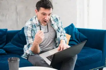Man in a casual plaid shirt sits on a blue couch, looking at a laptop with a focused expression, holding glasses in one hand. A coffee cup is on a table nearby.