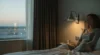 Woman sitting on a bed in a dimly lit room near a window with a glass of water on the nightstand and city lights outside.