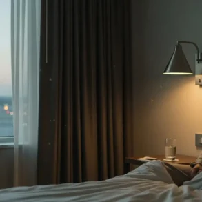 Woman sitting on a bed in a dimly lit room near a window with a glass of water on the nightstand and city lights outside.