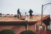 Roofers work on a shingled roof with stacks of new shingles, a chimney, and tools, while one worker ascends a ladder.
