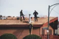 Roofers work on a shingled roof with stacks of new shingles, a chimney, and tools, while one worker ascends a ladder.