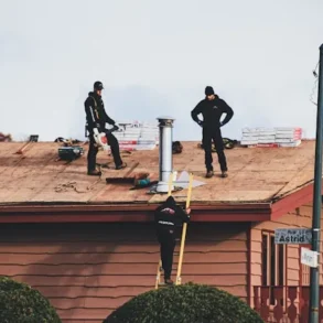 Roofers work on a shingled roof with stacks of new shingles, a chimney, and tools, while one worker ascends a ladder.