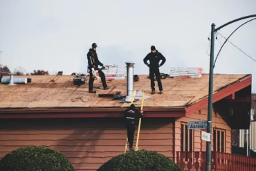 Roofers work on a shingled roof with stacks of new shingles, a chimney, and tools, while one worker ascends a ladder.
