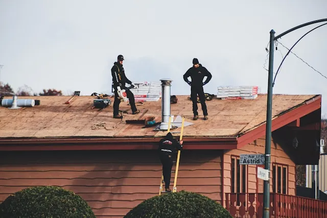 Roofers work on a shingled roof with stacks of new shingles, a chimney, and tools, while one worker ascends a ladder.