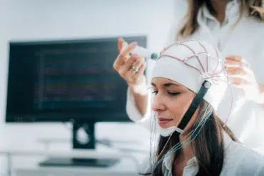 A woman wears an EEG cap connected with electrodes while another person adjusts it. A computer monitor with brainwave data is visible in the background.