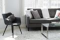Modern living room with gray sofa, black chair, glass coffee table, and patterned cushions on a light rug near large windows.